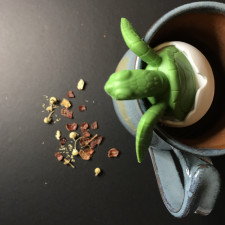 A turtle tea infuser resting over the lid of a hand-pottered turquoise mug with a chamomile loose leaf tea blend sitting on counter below.