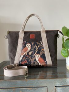 Handcrafted crossbody/tote bag with 2 owls in the front design. Made of brown waxed canvas, tan nylon webbing, and has 2 exterior pockets, 1 inner zippered pocket, 2 divided slip pockets inside, 1 key clasp, and an attachment to hold a water bottle.
